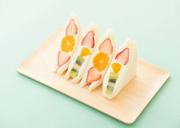 Fruit sandwiches filled with strawberries, kiwi, mango and oranges, set against a pale blue background.